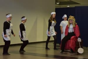 Mężczyzna w stroju króla z koroną na głowie siedzi na drewnianej skrzyni. Wokoło niego chodzą kobiety przebrane za pokojówki. W tle mężczyzna przebrany za kucharza.