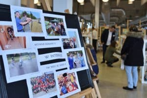 Na sztaludze jest kolaż różnych zdjęć z aktywności sportowych takich jak strzelanie z łuku, jazda na rowerze. W tle ludzie na korytarzu.