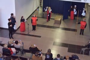 Ludzie stojący na scenie ubrani w czarno czerwone stroje. Jedna z kobiet stoi przy stojącym mikrofonie. Widownia siedzi na krzesłach.