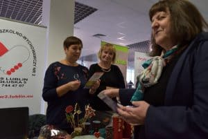 Kobieta uśmiecha się. W dłoniach trzyma ulotkę. W tle dwie uśmiechające się kobiety. Jedna z nich również w dłoniach trzyma ulotkę.