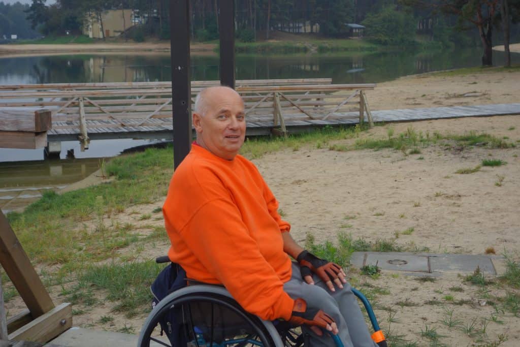 Mężczyzna na wózku inwalidzkim w pomarańczowej bluzie i skórzanych rękawiczkach pozuje bokiem do zdjęcia. w tle umocowany pomost nad akwenem wodnym.