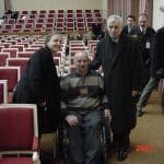 Kobieta i dwóch mężczyzn z czego jeden na wózku inwalidzkim pozują do zdjęcia w opustoszonej sali konferencyjnej. w tle osoby opuszczające salę. W prawym dolnym rogu czerwona data: 2002.1.22.