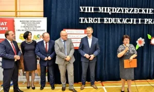 Na sali gimnastycznej stoi grupa osób- dwie kobiety i czterech mężczyzn. Kobieta po prawej stronie trzyma mikrofon blisko twarzy a w drugiej ręce ma teczkę.