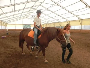 W stajni na koniu siedzi mężczyzna. Po lewej stronie na lince tego konia trzyma kobieta.