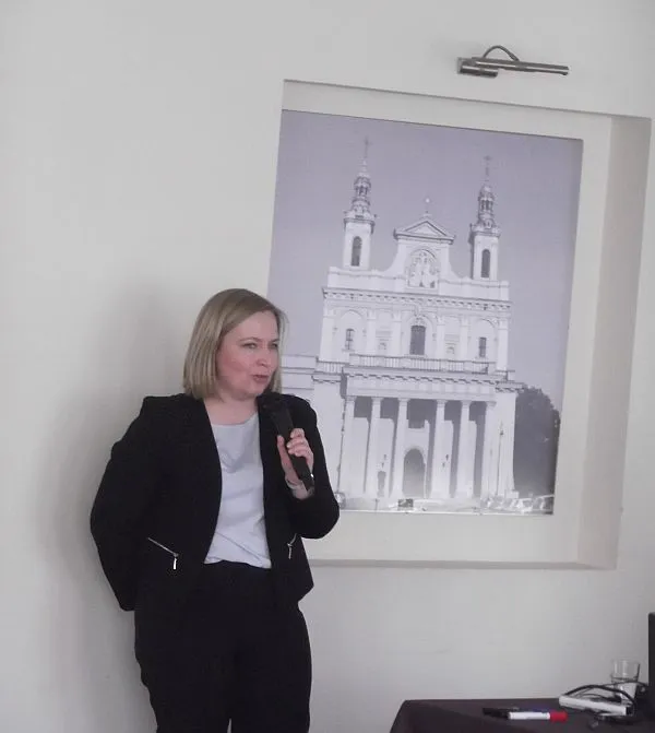 Kobieta opiera się o ścianę. Trzyma w dłoni mikrofon. W tle wisi obraz na ścianie.