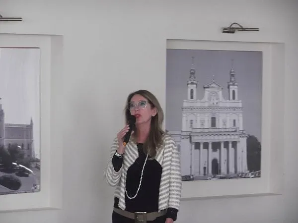 Kobieta stoi i trzyma mikrofon w dłoni. Za nią na ścianie wisi obraz.