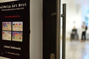 Po lewej stronie na ścianie wisi plakat “Galeria Art Brut - Centrum Kultury w Lublinie”. Obok zaraz są przeszklone drzwi przez które widać dwie osoby w pomieszczeniu obok.