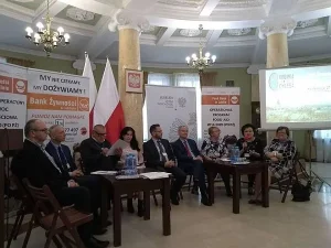 Kobiety i mężczyźni siedzą na sali. Za nimi stoją banery z napisami m.in. “ My nie czekamy my dożywiamy!”. Trzeci mężczyzna od lewej trzyma w rękach kartki papieru.