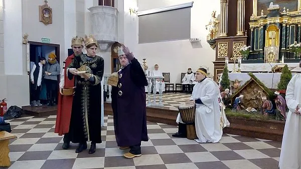 Ludzie przebrani za trzech króli stoją przed ołtarzem na środku w kościele. W tle osoba przebrana za anioła ma bębenek między nogami.