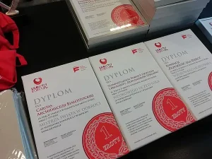 Dyplomy leżą na stole. W dolnym prawym rogu jest grafika czerwonej złotówki.