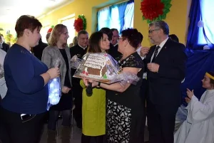 Na zdjęciu jest grupka ludzi. Dwie Panie stoją naprzeciwko siebie w małej odległości i patrzą w swoim kierunku. Jedna z tych Pań trzyma w lewej ręce prezent.