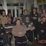 Siedmiu mężczyzn na wózkach inwalidzkich, za nimi siedem stojących osób, pozują do aparatu. Sześcioro z nich ma na ubraniach żółte etykietki imienne.