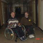wóch mężczyzn z wąsami na wózkach inwalidzkich pozują z rękami w geście przywitania na długim korytarzu. w tle stoją dwie osoby przy drzwiach wyjściowych. W prawym dolnym rogu czerwona data: 2001.10.26.