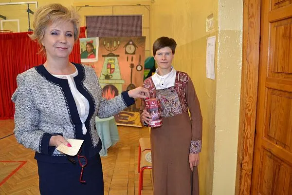 Na pierwszym planie znajdują się dwie kobiety. Jedna z nich trzyma w ręku puszkę, druga trzyma karteczkę.