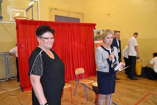 Na pierwszym planie są dwie kobiety. JEdna z nich trzyma w prawym ręku mikrofon przy ustach. Obie patrzą się wprost przed siebie. W tle widać mężczyzn.