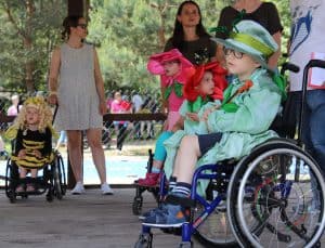 Czworo dzieci w przebraniach, z czego troje jest na wózkach inwalidzkich. Za nimi stoją opiekunowie