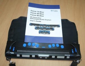 Na stole w kolorze drewna umieszczony jest w futerale czarny monitor brajlowski z niebieskimi klawiszami. Nad monitorem leży instrukcja obsługi sprzętu.