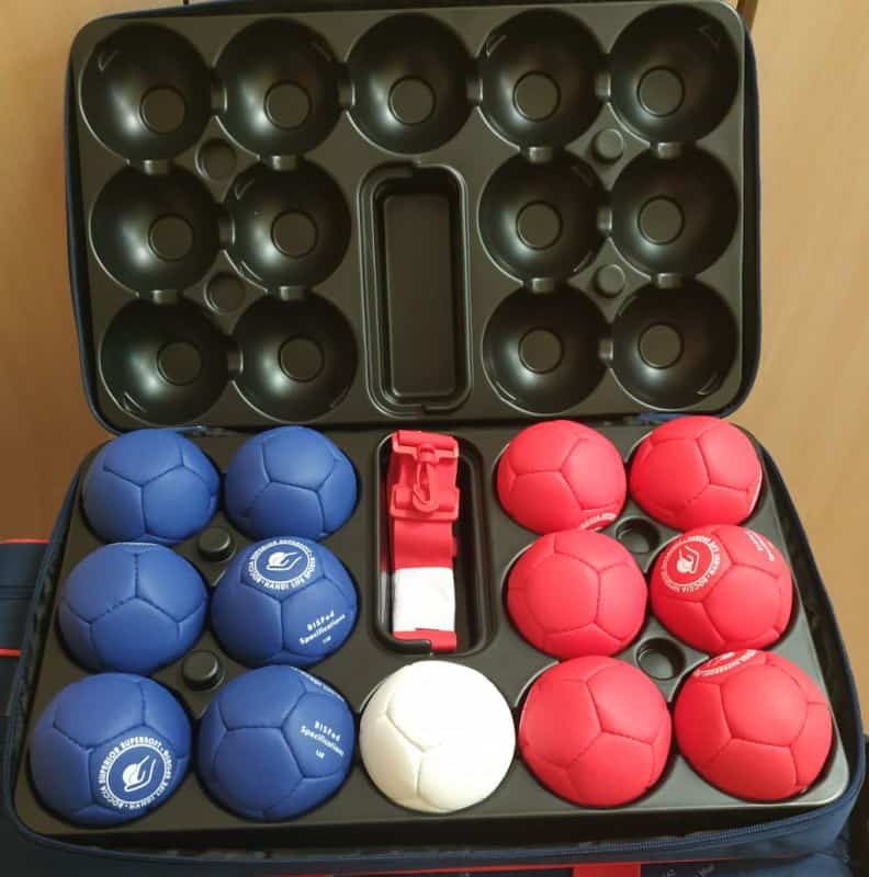 Otwarta walizka, w której znajduje się sprzęt do gry boccia. W walizce po prawej stronie w dwóch kolumnach znajduje się sześć czerwonych bil, po środku w jednej kolumnie znajduje się jedna biała bila oraz pasek do walizki, zaś po lewej stronie w dwóch kolumnach - sześć niebieskich bil.