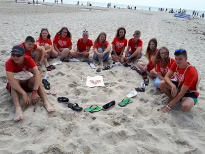 Ludzie siedzący w półokręgu na plaży. W środku ich półokręgu ułożone są buty w kształcie serca w którym leży plażowy plecach z napisem caritas układającym się w kształt serca.