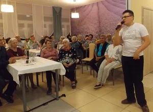 Mężczyzna stoi na środku sali. Trzyma w dłoni mikrofon blisko ust, drugą dłoń ma włożoną do kieszeni od spodni. W tle starsi ludzie siedzący na krzesłach. Twarze mają skierowane w stronę stojącego mężczyzny w mikrofonem.