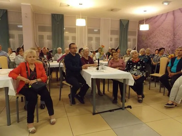 Osoby w wieku starszym siedzą przy stole w sali. Ich twarze są skierowane na wprost do zdjęcia. Niektórzy opierają się rękoma o stół.