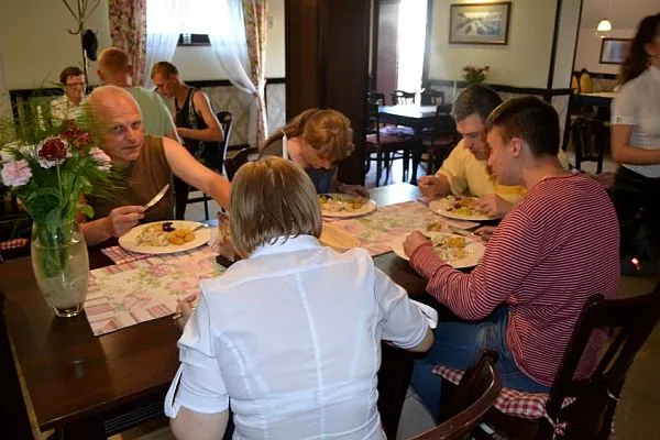 Dwie kobiety i trzech mężczyzn spożywają posiłek przy jednym stole. Mężczyzna patrzy w obiektyw, trzymając w ręku nóż. Pozostali jedzą.