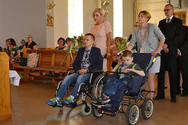 Chłopiec i mężczyzna są na wózkach inwalidzkich. Mężczyzna patrzy przed siebie, natomiast chłopiec ma spuszczony wzrok. Za nimi stoją dwie kobiety, trzymające wózki. W tle stoją i siedzą w ławkach kościelnych inni ludzie
