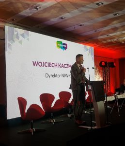 Mężczyzna w garniturze stojący na scenie przy mównicy. Przy jego ustach mikrofony. W tle za nim krzesła.