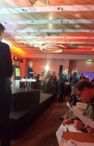 Kobieta stojąca na scenie z mikrofonem w dłoni. Poniżej sceny dwóch mężczyzn w uścisku dłoni. w oddali reporterzy i światło kamer.
