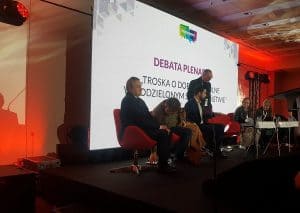Ludzie siedzący w fotelach na scenie. W tle ekran multimedialny wyświetlający napis - debata plenarna.