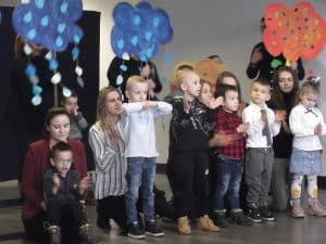 Kobiety stojące na scenie z dziećmi. Dzieci klaszczą w swoje dłonie. Nad głowami dzieci trzy kobiety trzymają malowane chmurki z wyciętymi kroplami deszczu.