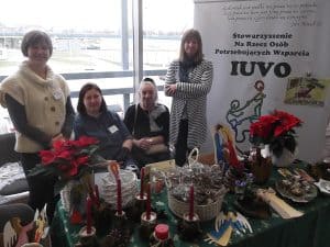 Uśmiechające się do zdjęcia kobiety stojące przy stole rękodzieł. Na stole świece woskowe, kwiaty gwiazdy betlejemskiej.