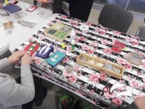 Dłonie osoby przyklejają karteczkę do ręcznie wykonanej kartki bożonarodzeniowej. Na stole otwarty sztyft kleju oraz pudełeczka z ozdobami.