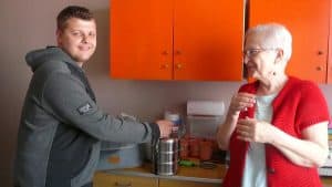 Mężczyzna trzyma w dłoni garnek aluminiowy. Jego twarz skierowana jest na wprost do zdjecia, uśmiecha sie. W jego kierunku obraca sie starsza kobieta po prawej.