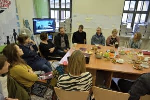 Ludzie siedzą przy stole. Stół jest jest zastawiony jedzeniem. W tle telewizor.