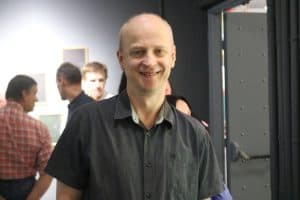 Mężczyzna pozuje do zdjęcia. Stoi tuż przed obiektywem i uśmiecha się. Za nim są ludzie.