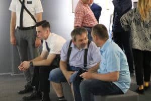 Grupa osób jest w pomieszczeniu. Trzech mężczyzn siedzi na pufach. Mężczyzna pośrodku jest odwrócony twarzą do mężczyzny po prawej stronie. Wszyscy są bokiem do obiektywu.