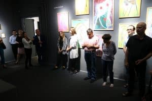 W sali na ścianach wiszą obrazy. Ludzie stoją tyłem do nich obok siebie. Prawa część sali jest bardziej oświetlona niż lewa.