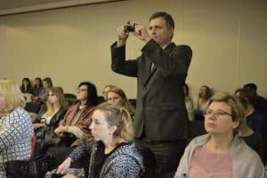 Osoby siedzą na widowni. Mężczyzna stoi pośród kobiet i trzyma aparat fotograficzny w rękach. Wszyscy patrzą przed siebie.