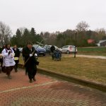 Kondukt pogrzebowy idzie chodnikiem. Obok trzy osoby na wózkach inwalidzkich poruszają się po trawniku. W tle parking z autami.