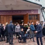 Przed głównym wejściem kościoła oczekują zgromadzeni uczestnicy ceremonii pogrzebowej, w tym trzy osoby na wóżkach inwalidzkich. Wśrod nich dwie przytulające się kobiety.