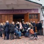 Zgromadzeni uczestnicy ceremonii pogrzebowej, w tym trzy osoby na wóżkach inwalidzkich przed głównym wejściem do kościoła. Nad drzwiami kościoła napis: EUCHARYSTIA DAJE ŻYCIE!