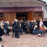 Po lewej stronie trzech panów z konduktu przygotowujących do przeniesienia urny i wiązanek kwiatów z karawanu na Mszę Świętą do kościoła. Na środku zgromadzeni uczestnicy ceremonii pogrzebowej, w tym trzy osoby na wóżkach inwalidzkich przed głównym wejściem do kościoła.