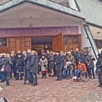 Zgromadzeni uczestnicy ceremonii pogrzebowej przed głównym wejściem do kościoła. Wsród nich są osoby na wózkach inwalidzkich i jedna starsza pani o kuli. Po lewej stronie część otwartego karawanu pogrzebowego z widoczną urną i wiązankami kwiatów.