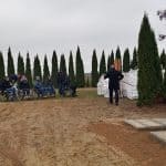 Kobieta i czterech mężczyzn na wózkach inwalidzkich z dwiema osobami asystującymi na tle szpalerów z tui. Przed nimi po prawej stronie przygotowane groby do pochówku.