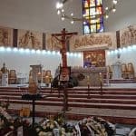 Na pierwszym planie urna w kolorze beżowym, obok portret mężczyzny. Przed urną tabliczka nagrobna: Adam Orzeł a wokół wieńce i wiązanki pogrzebowe. W tle ksiądz przed ołtarzem przewracający mszał.