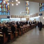 Uczestnicy ceremonii siedzący w ławkach tyłem bocznym do obiektywu. W tle ołtarz i kolorowe witraże na oknach.