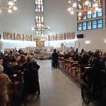 Widok na główną nawę do ołtarza. Po bokach ławki z uczetnikami, przy nich osoby siedzące na wózkach inwalidzkich. W tle urna z wiązankami i ołtarz.