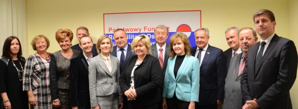 Zdjęcie grupowe: Rada Nadzorcza i Zarząd Państwowego Funduszu Rehabilitacji Osób Niepełnosprawnych. W tle na ścianie duże logo PFRON