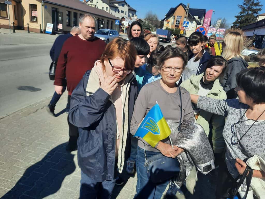 Grupa osób stojących na ulicy, jedna z nich - kobieta trzyma w ręku małą flagę ukraińską. W tle budynki.